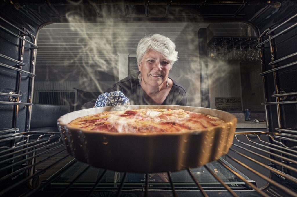 visão de dentro de um forno de cozinha, com uma torta de maçã em primeiro plano e uma senhora idosa ao fundo, pegando a torta.